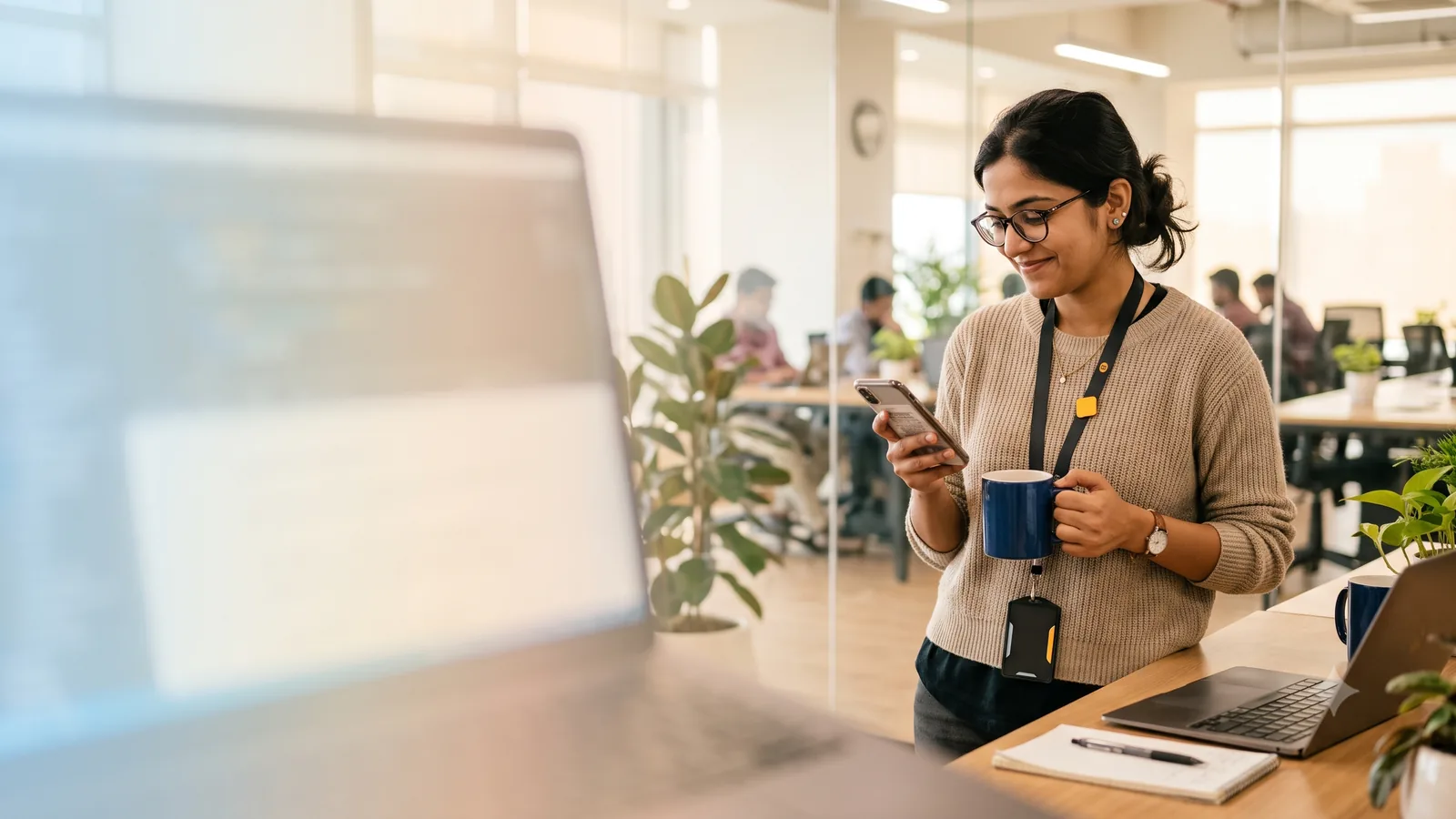An IT professional in a Mumbai tech-corridor office glancing at her phone with a bemused half-smile, laptop blurred in the foreground.